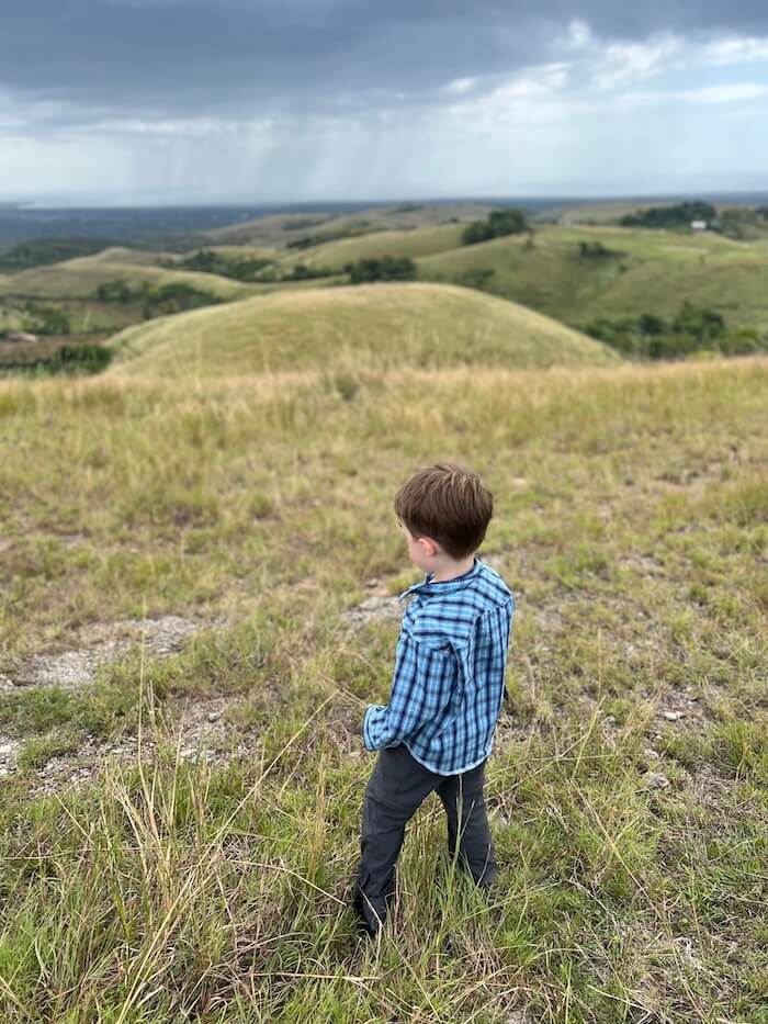 A young boy in a blue plaid shirt standing in a grassy field, looking out over rolling green hills under a vast, overcast sky.