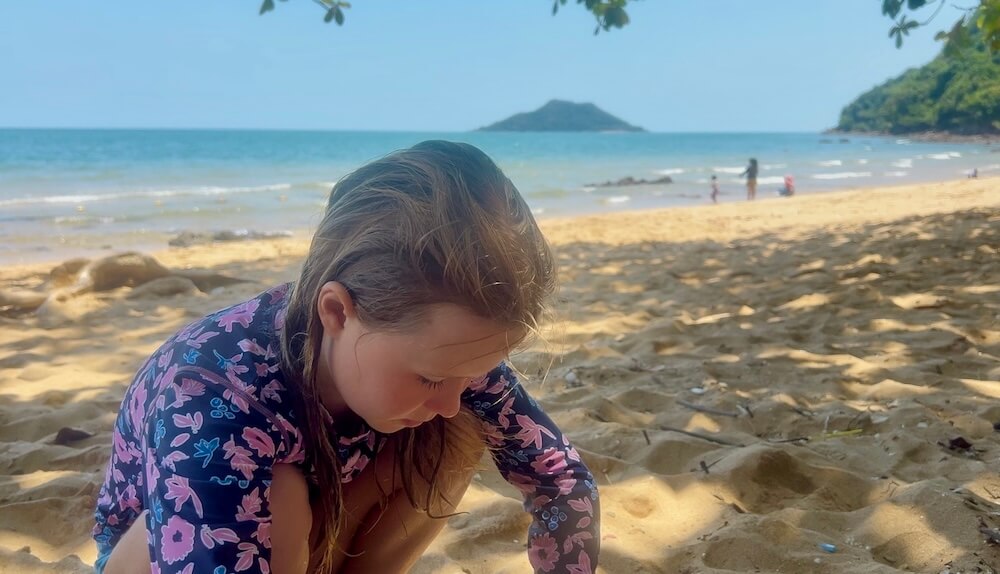 A young girl playing in the red sand under the shade of broad branching trees at Ao Krathing beach in Chanthaburi.