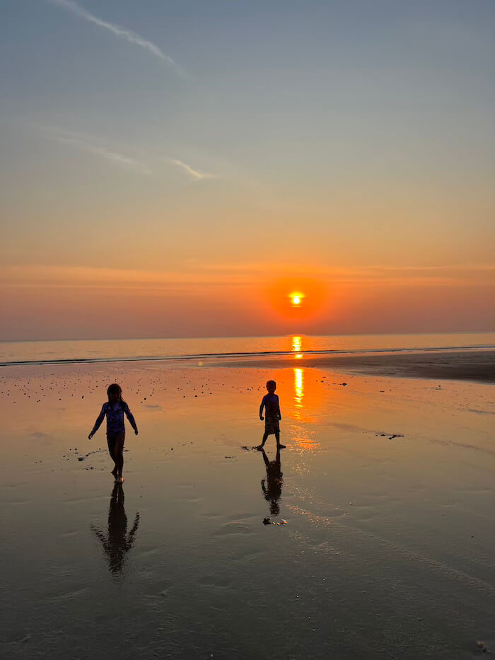 Sunset Magic on Chakphong Beach Two young children silhouetted against a vibrant orange and pink sunset, walking along the reflective, shallow water's edge of a quiet Chakphong Beach in Rayong, Thailand.