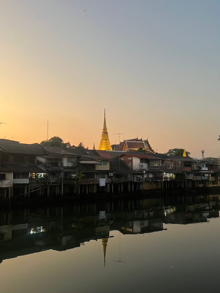 A wide view of the Chanthaburi River at sunset with old wooden houses on stilts and a large golden Buddhist chedi reflecting in the glassy water.