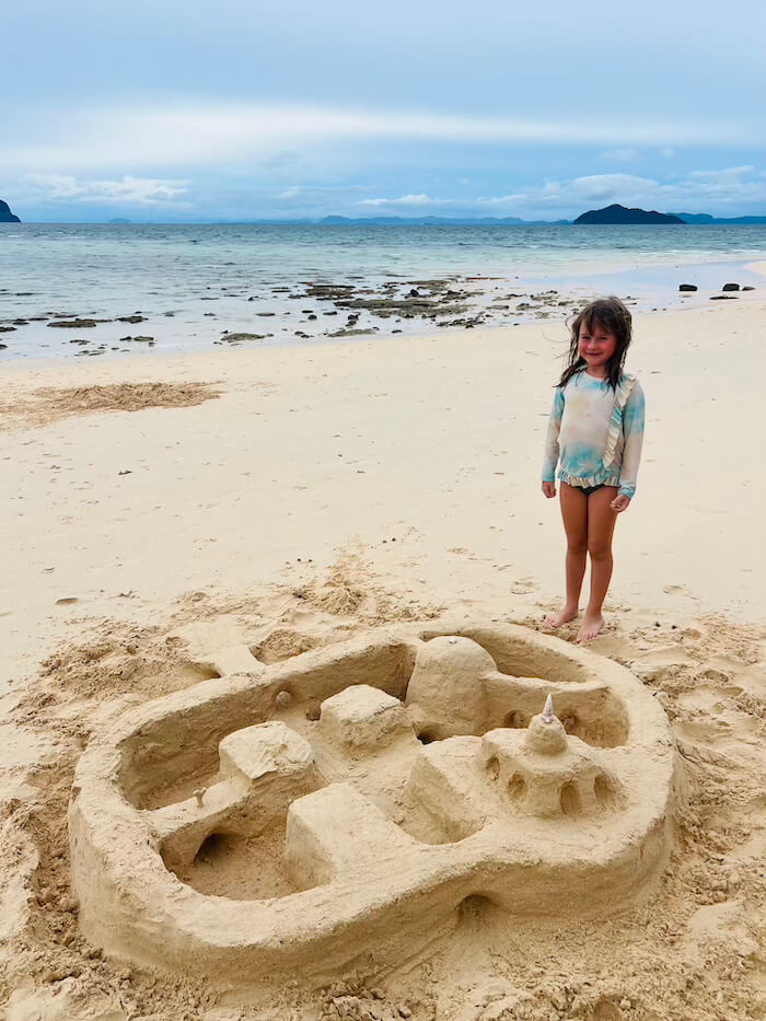 Monumental Sandcastles on Bulon A young girl standing proudly next to a large, intricately designed sandcastle on a wide, white sand beach at Koh Bulon Lae.
