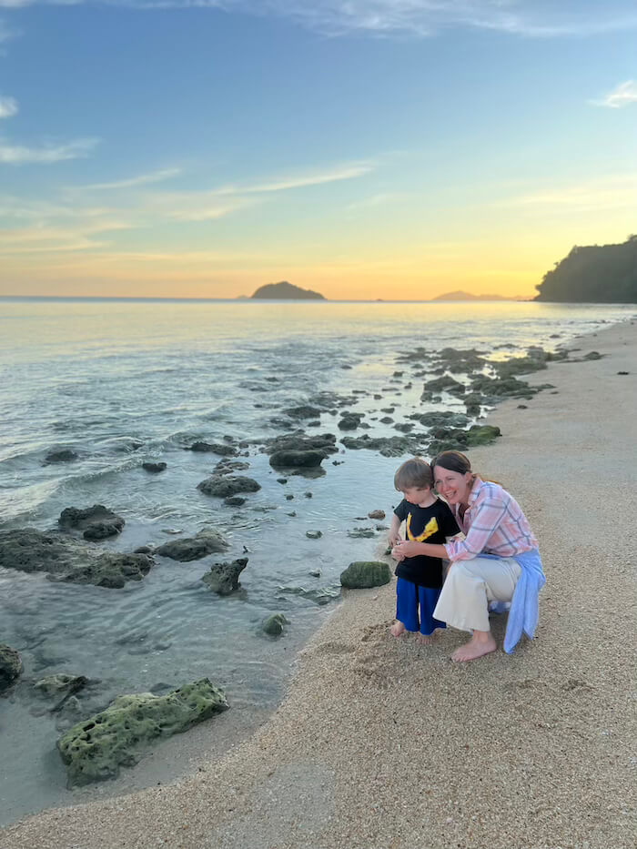 Shoreline Discoveries A mother and young son crouched on a sandy beach at Koh Bulon Lae, looking at rocks and shells at the water's edge during sunset.