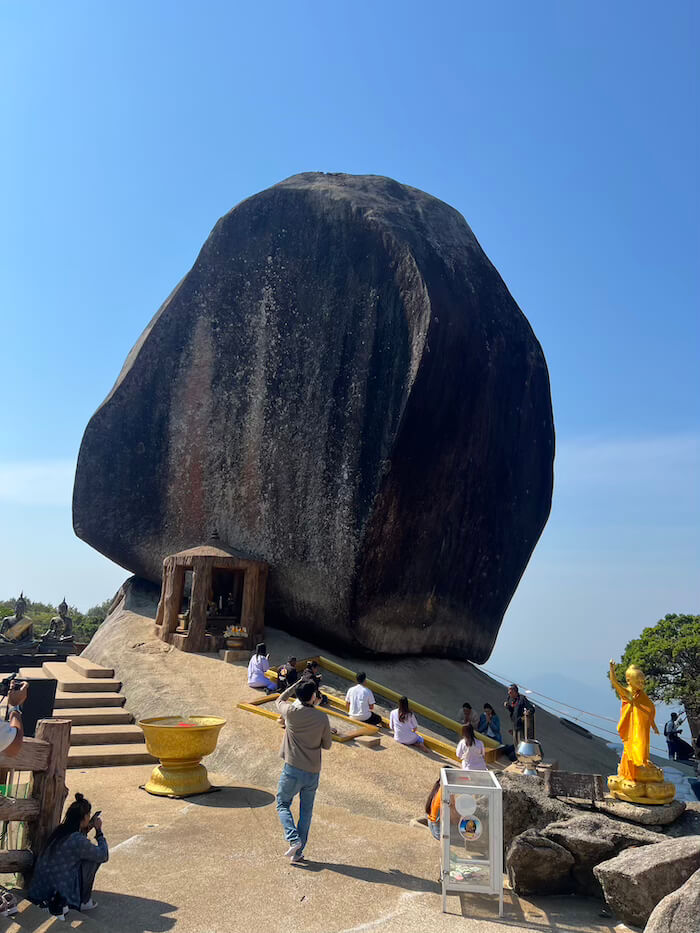 A massive, dark granite boulder perched precariously on a mountain cliff at Khao Khitchakut, with pilgrims praying at its base under a bright blue sky.