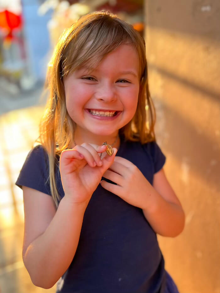 A young blonde girl smiling broadly while holding up a small golden religious medal on a necklace in the golden afternoon sun.