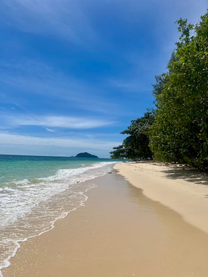 koh-bulone-lae-pristine-beach A wide, empty white sand beach on Koh Bulon Lae with turquoise water on the left and lush green trees on the right under a bright blue sky.