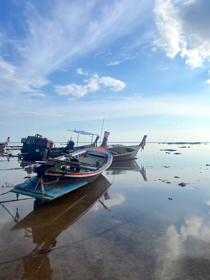 Traditional Thai longtail boats anchored in shallow, mirror-like water during low tide on Koh Libong, with clouds reflected in the sea.