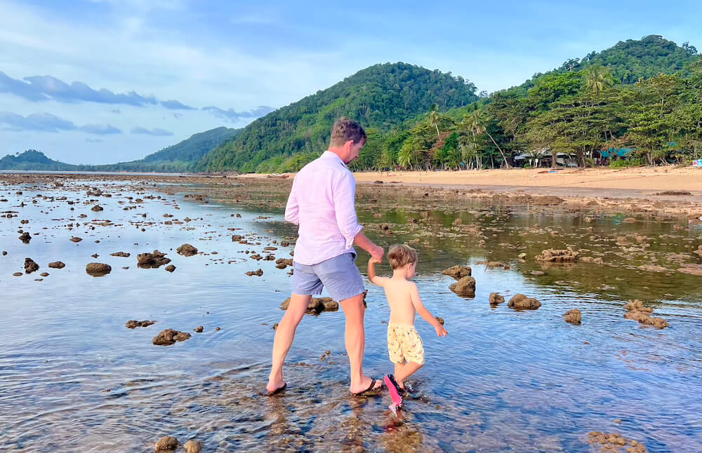 Walking the Libong Seabed A man and a small child walking hand-in-hand across the exposed seabed and coral flats of Koh Libong during low tide, with lush green hills in the background.