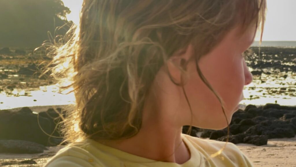 Golden Hour on the Tidal Flats Close-up of a young girl's profile with sunlight catching her hair at sunset on Koh Libong, with the rocky shoreline and sea in the blurred background.