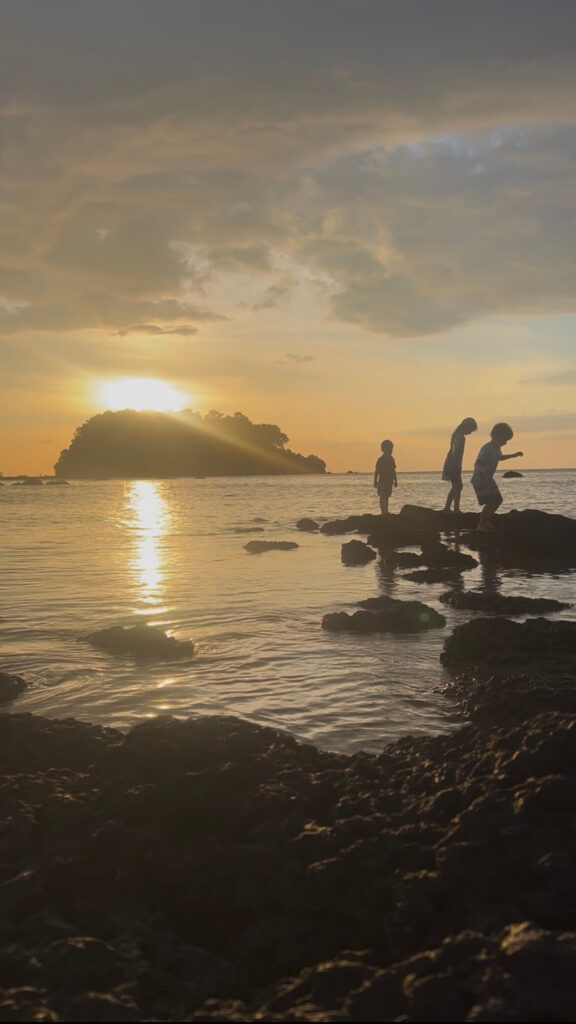 Sunset Explorations on Koh Libong Silhouette of three children playing on rocks in the sea at sunset on Koh Libong, with the jungle-covered Koh Nu island in the background.