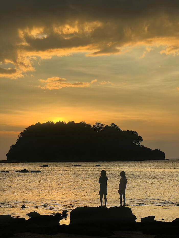 Golden Hour at Mouse Island Silhouette of two children standing on rocks at dusk, looking toward the dark shape of Koh Nu island under a vibrant orange and yellow sunset sky.