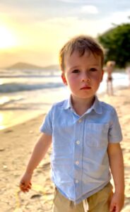 A young boy in a blue button-down shirt standing on a sandy beach at sunset in Rayong, Thailand.