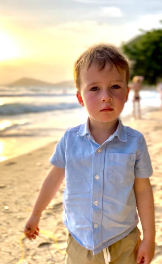 A young boy in a blue button-down shirt standing on a sandy beach at sunset in Rayong, Thailand.
