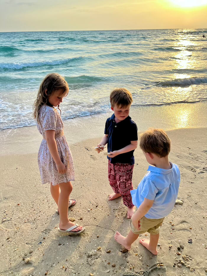 Golden Hour on Ao Nang Beach , Koh Samet Three young children—a girl and two boys—examining shells or treasures on a sandy Koh Samet beach at sunset, with gentle turquoise waves in the background