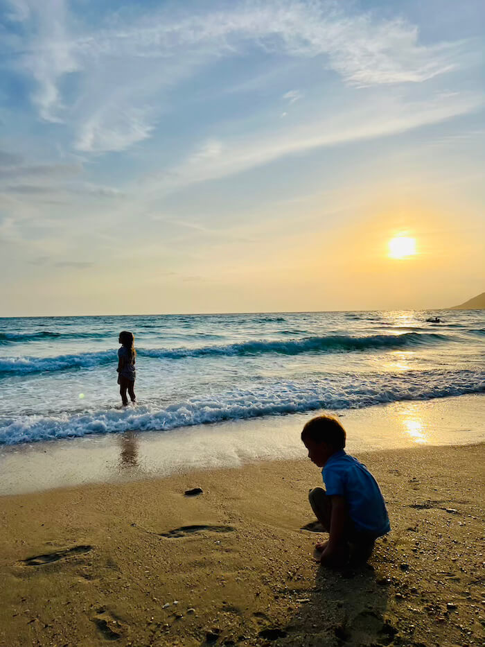 Quiet Evenings on Koh Samet Silhouettes of two children playing on a Koh Samet beach at sunset, with one child standing in the shallow turquoise surf and another squatting on the sand in the foreground.