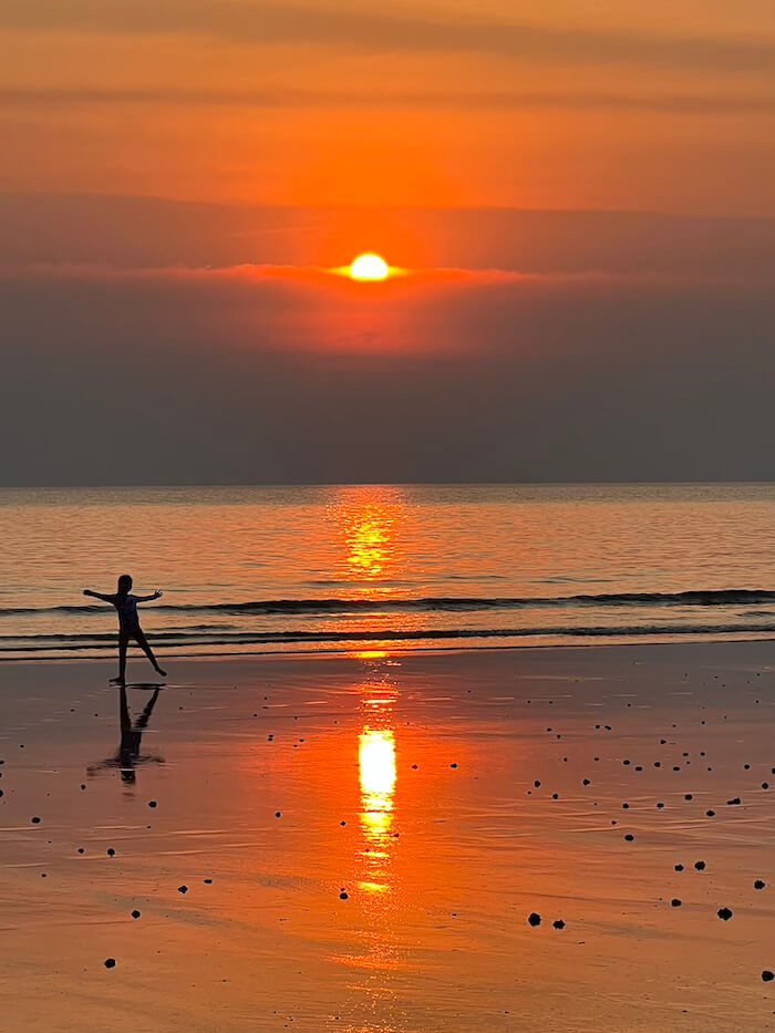 Golden Hour at Laem Mae Phim A young child silhouetted with arms outstretched against an intense orange sunset on the reflective, wet sand of Laem Mae Phim Beach in Rayong, Thailand