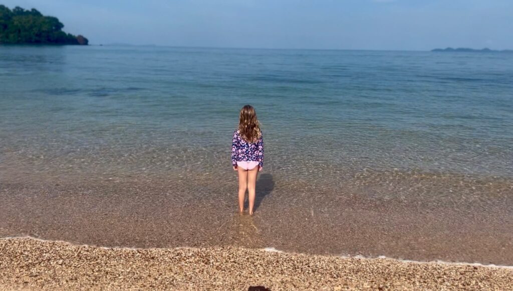The Golden Shores of Koh Libong A young girl in a floral swim shirt standing at the water's edge on the golden, pebbly sand of Lang Khao Beach, Koh Libong, looking out at the calm turquoise sea.