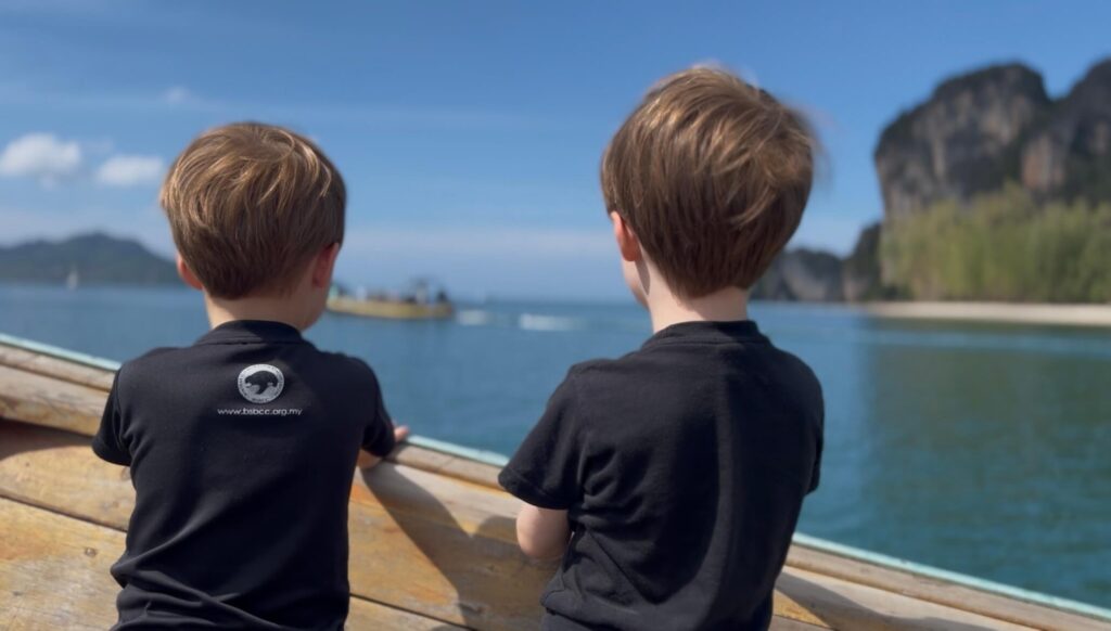longtail-hat-yao-banphrao-koh-libong Two young boys in black t-shirts looking out from a longtail boat toward the towering limestone cliffs of Koh Lao Liang, Thailand, on a bright sunny day.