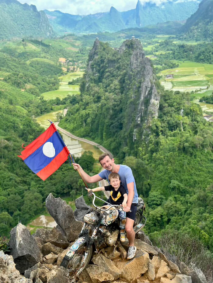 A man and a young child sit on a vintage motorcycle bolted to the edge of a limestone cliff at Nam Xay Viewpoint, Vang Vieng, holding the Laotian flag overlooking a valley.