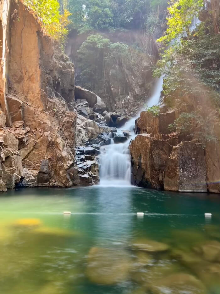A multi-tiered waterfall cascading into a clear, emerald-green pool surrounded by tall rock cliffs and lush jungle in Namtok Phlio National Park.