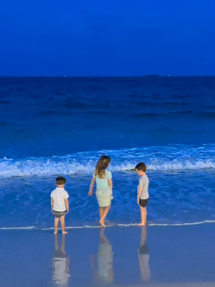 he Magic of the Blue Hour Three young children standing at the water's edge of a dark blue sea during the deep blue hour, their reflections visible in the wet sand under a twilight sky.