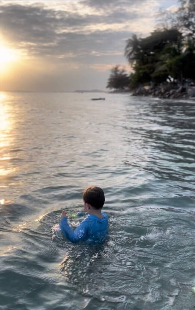 An Evening Dip in Rayong A young boy in a blue swim shirt swimming in the gentle waves of the Rayong coast at sunset, with the sun reflecting off the water