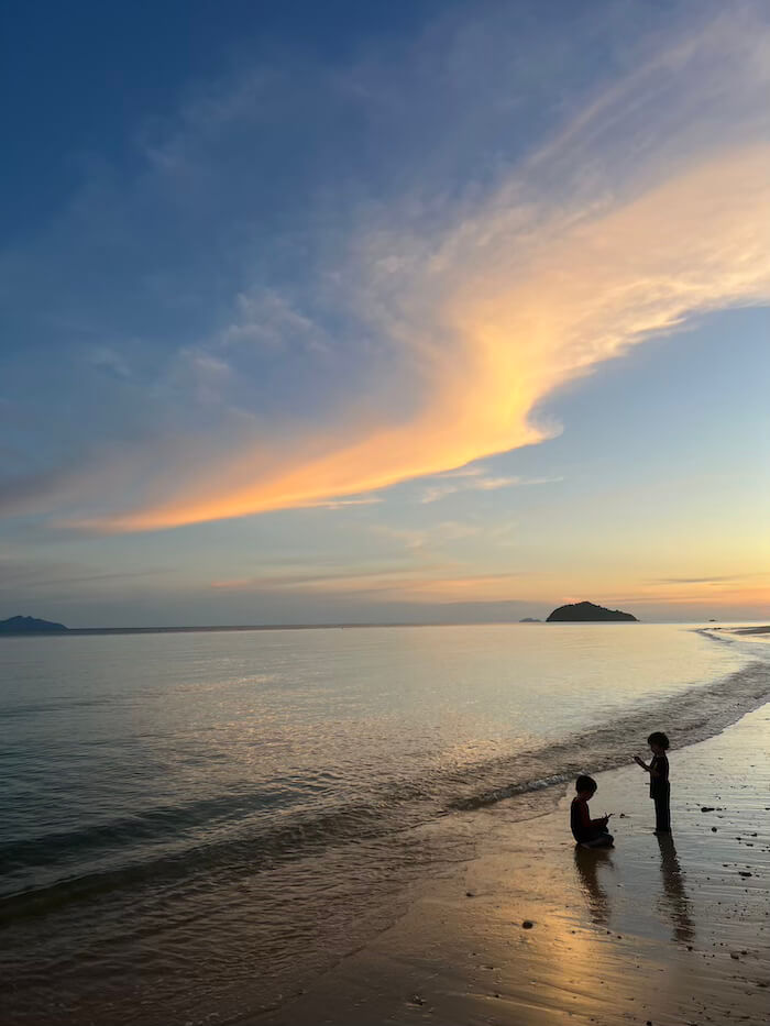 Golden Hour on School Beach Two young children playing at the water's edge on a sandy beach during a vibrant orange and blue sunset on Koh Bulon Lae.