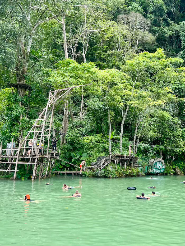 A view of Blue Lagoon 3 in Vang Vieng, Laos, showing the turquoise-green water filled with swimmers and people in inner tubes, set against a backdrop of dense jungle and a wooden jumping structure.