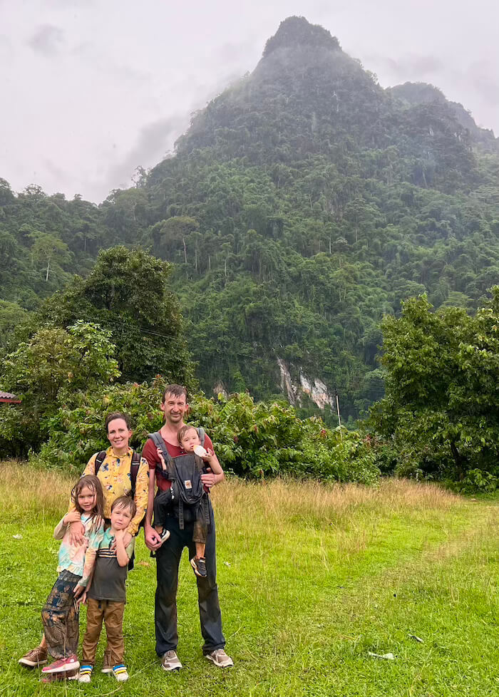 A family of five—two parents and three young children—standing in a grassy field, looking tired and covered in mud after a jungle hike in Vang Vieng, Laos, with a large jungle-covered mountain in the background.