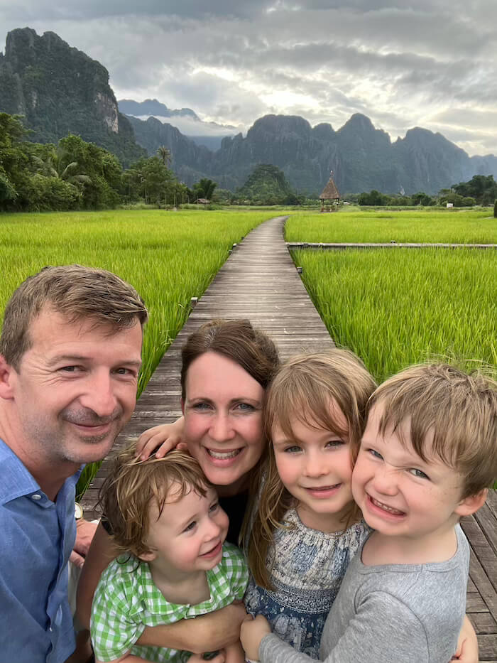 A smiling family of five—two parents and three young children—posing for a selfie on a wooden boardwalk in the middle of lush green rice fields in Vang Vieng, Laos.