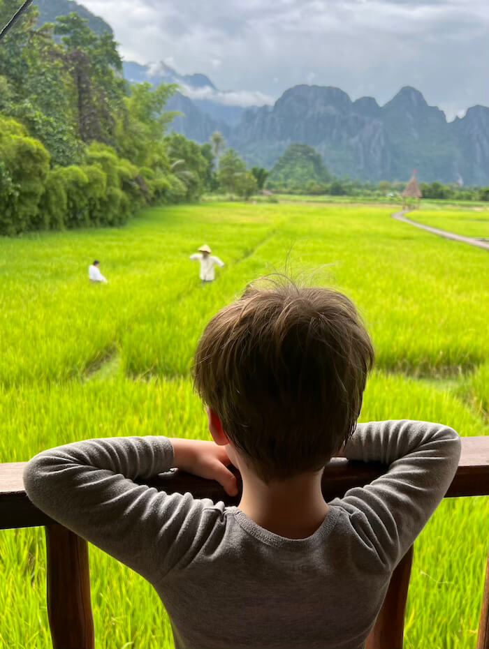 A young boy looks out from a wooden balcony over vibrant green rice paddies and limestone mountains in Vang Vieng, Laos.