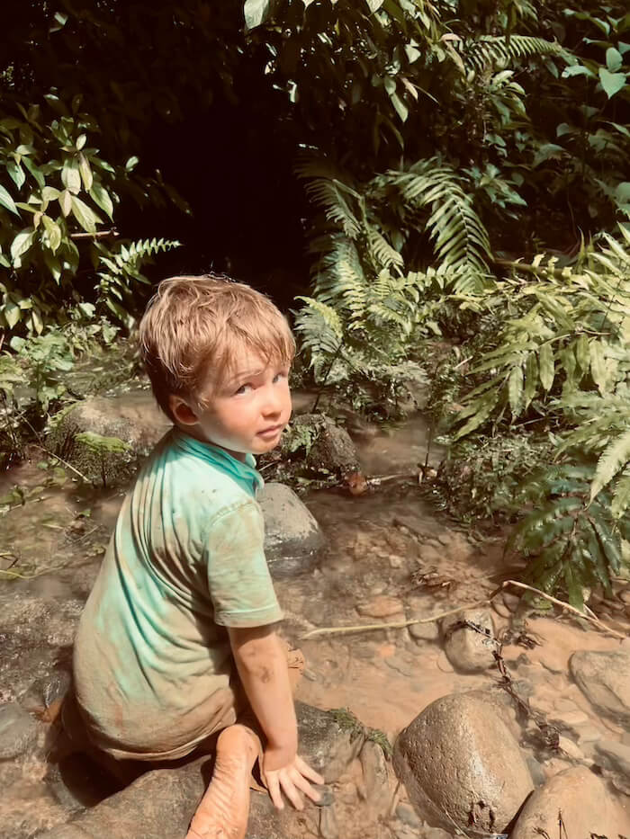 A young boy, covered in mud from a hike, sits by a shallow jungle stream in Vang Vieng, Laos, surrounded by lush green ferns.