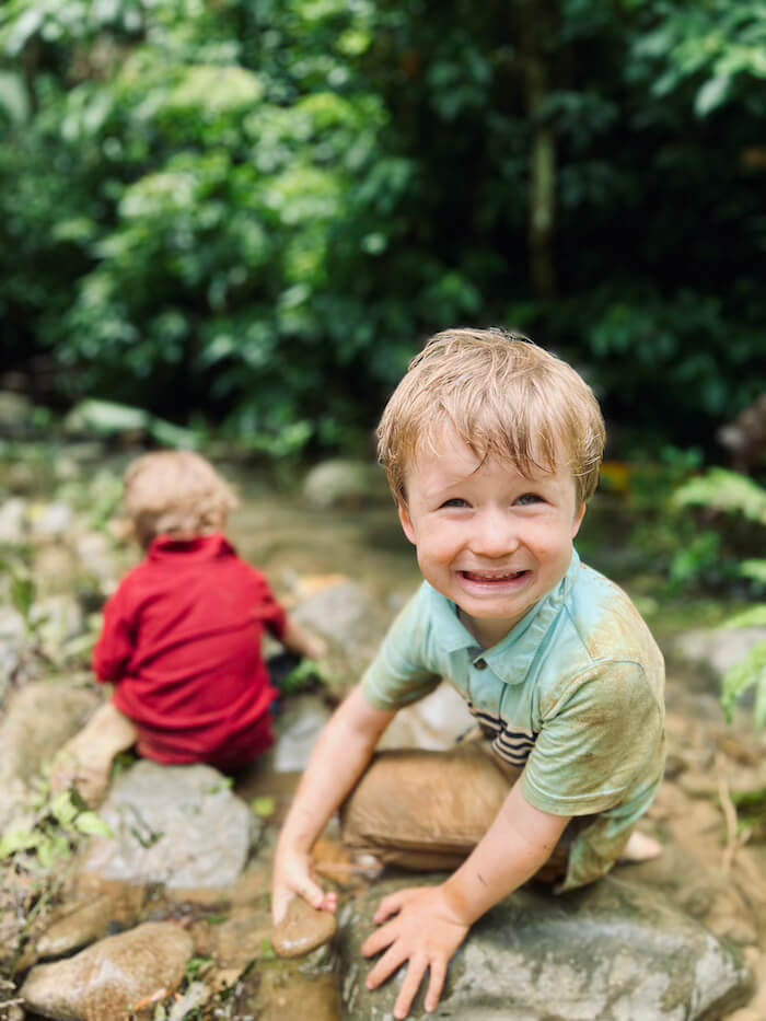 A young boy with a mud-stained teal shirt smiles broadly while playing on rocks in a jungle stream in Vang Vieng, Laos, as another child in a red shirt sits in the background.