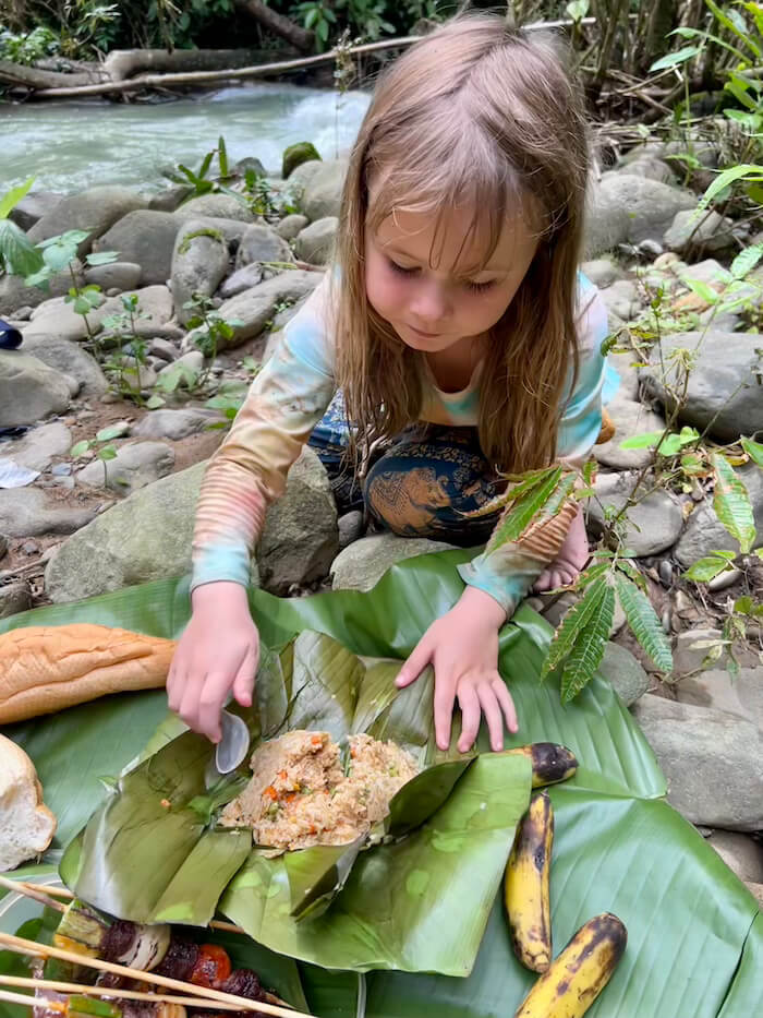 A young girl sitting on river rocks in Vang Vieng, Laos, eating fried rice served on a large green banana leaf with bread and grilled skewers.