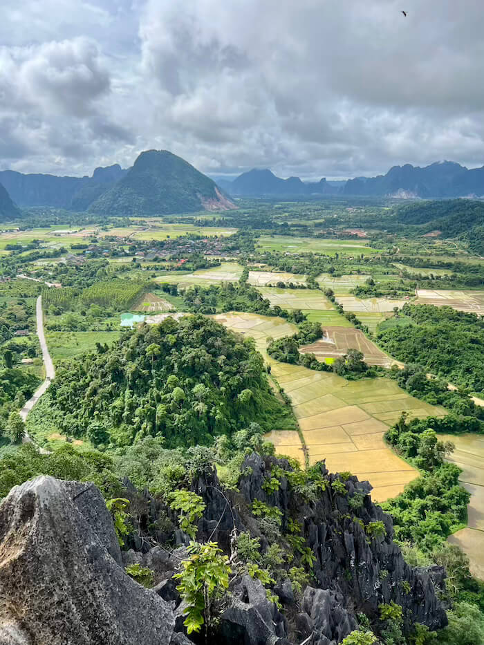 A high-angle panoramic view of the Vang Vieng valley from Nam Xay Viewpoint, showing flooded rice paddies, limestone karsts, and a winding road under a cloudy sky.