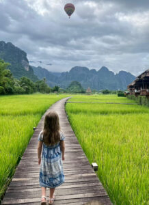 A young girl in a blue dress walks along a wooden boardwalk through lush green rice fields in Vang Vieng, Laos, with limestone mountains and a hot air balloon in the background.