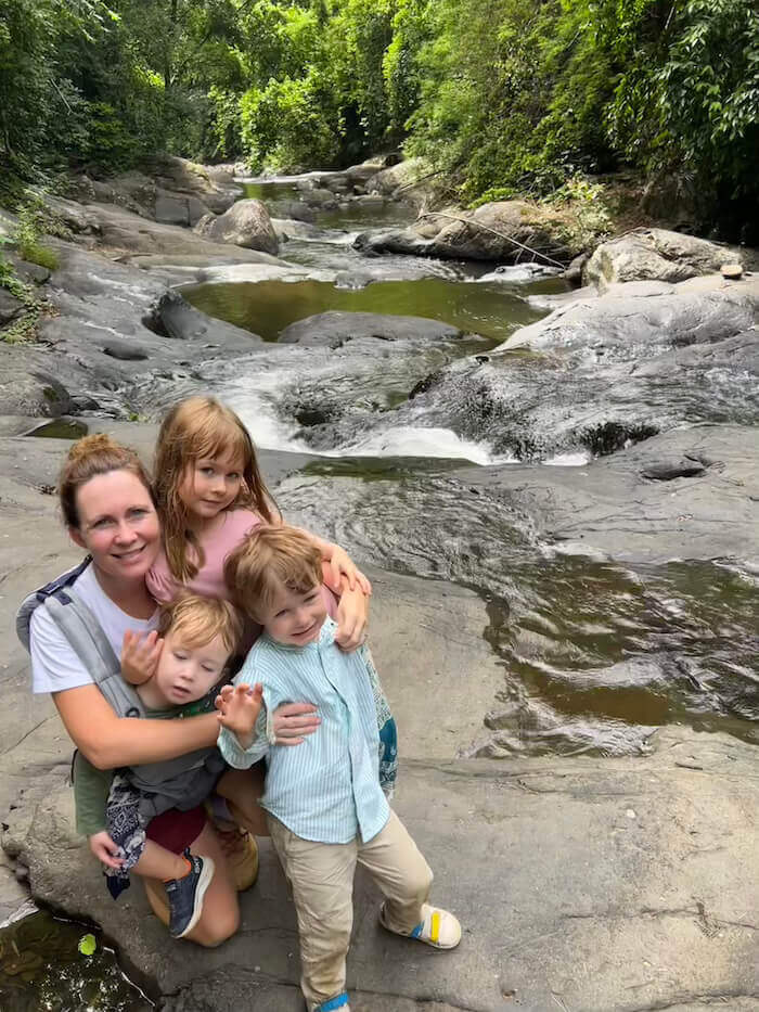 A mother and three young children smiling together on a large rock overlooking the flowing streams and jungle canopy of Pala-U Waterfall.