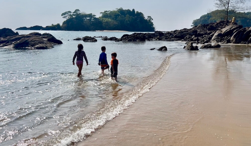 Three children wading into the shallow tide on a secluded beach with rocky outcrops and offshore islands in Koh Phra Thong, Thailand.