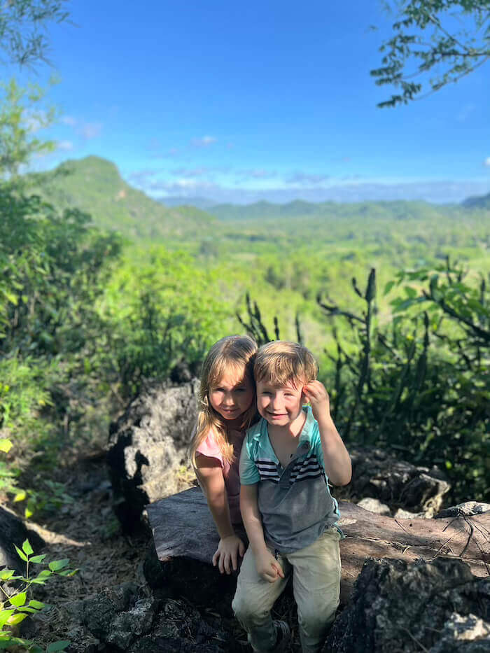 Two young children sitting on a rocky outcrop with a vast, lush green valley and rolling hills of Phetchaburi in the background under a clear blue sky.