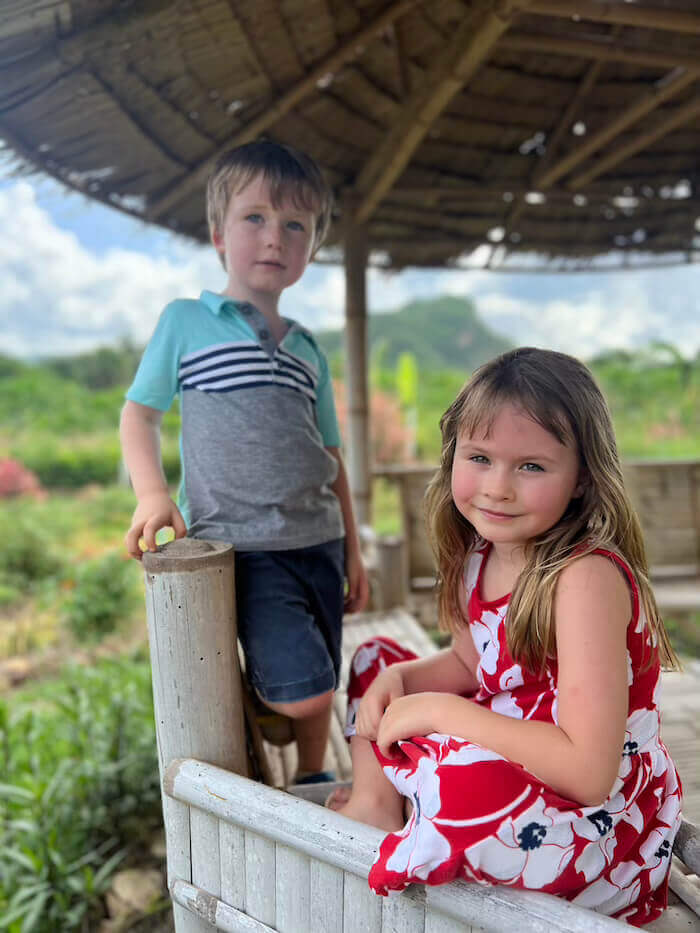 Two young children sitting together in a rustic bamboo gazebo at Baan Maka Nature Lodge, with lush green hills and a bright sky in the background.