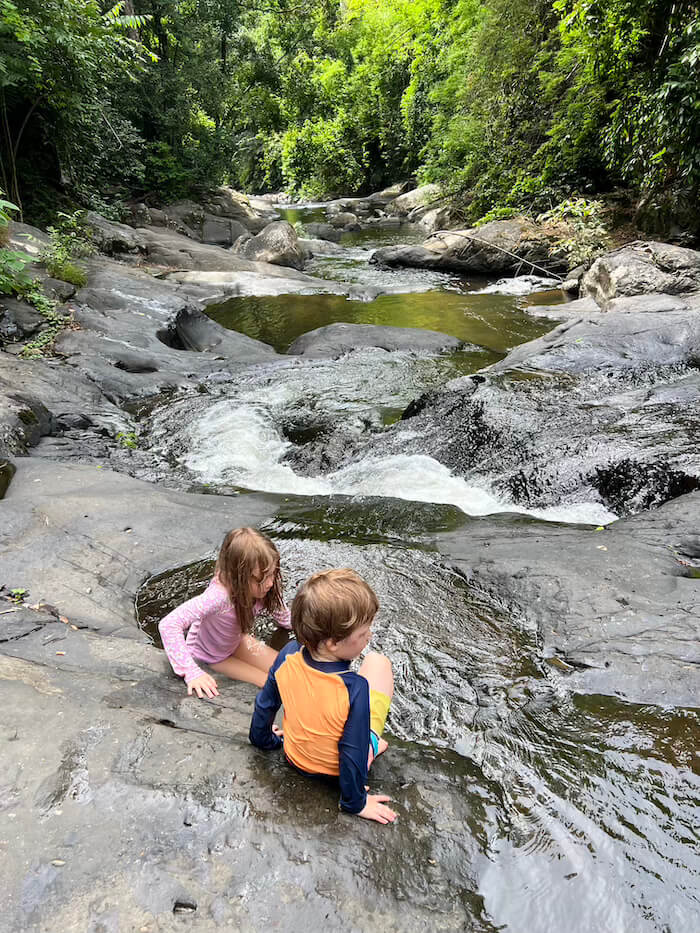 Two young children in swim shirts sitting and playing in shallow, clear rock pools at Pala-U Waterfall, surrounded by dense jungle.
