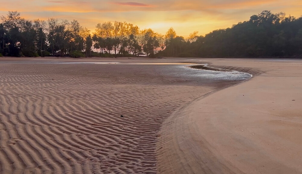 Ornate sand ripples created by the receding tide on a wide beach at sunset, with a golden sky and silhouetted palm trees in the background on Koh Phra Thong.