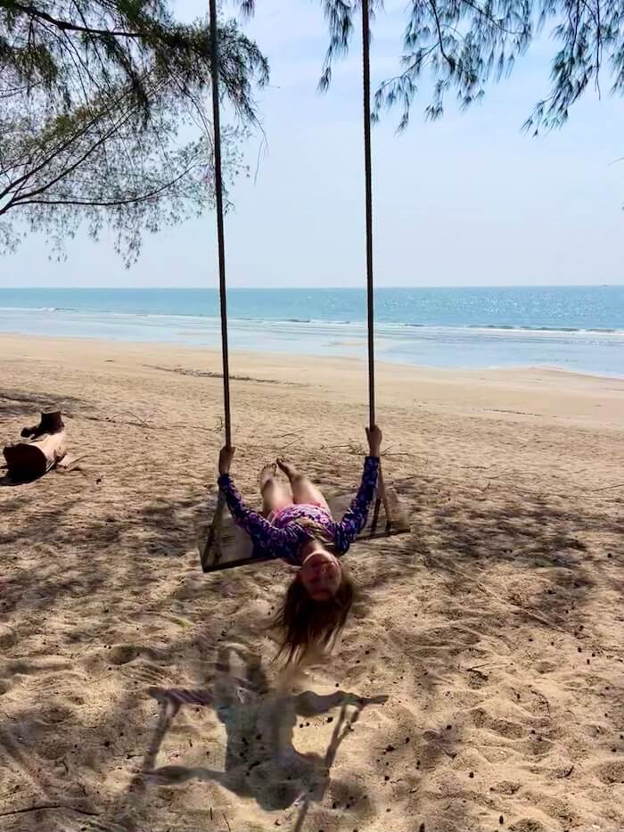 A young girl leaning back on a wooden rope swing hanging from a tree on a quiet, sandy beach with the turquoise Andaman Sea in the background.