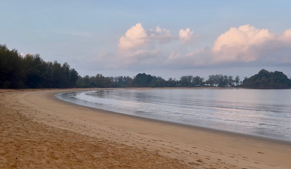 A wide, tranquil golden sand beach on Koh Phra Thong at sunrise, with soft pink clouds reflected in the still, mirror-like water and pine trees lining the shore.