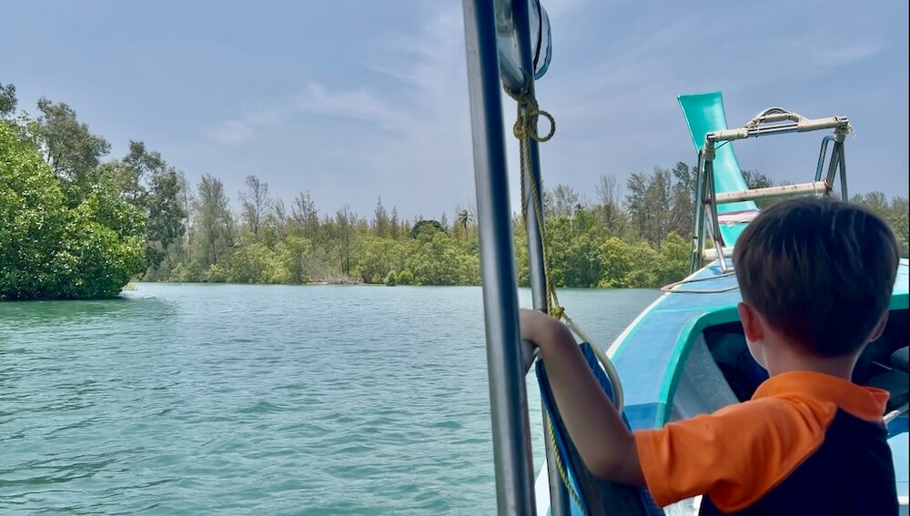 A young boy in an orange shirt looks out from a longtail boat toward the lush mangrove-lined coast of Phang Nag en route to Koh Phra Thong, Thailand.