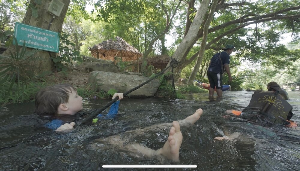 A young boy in a life jacket floating in the Phetchaburi River, holding onto a rope, with the lush riverside forest and bamboo huts of Kenaray Farmstay in the background.