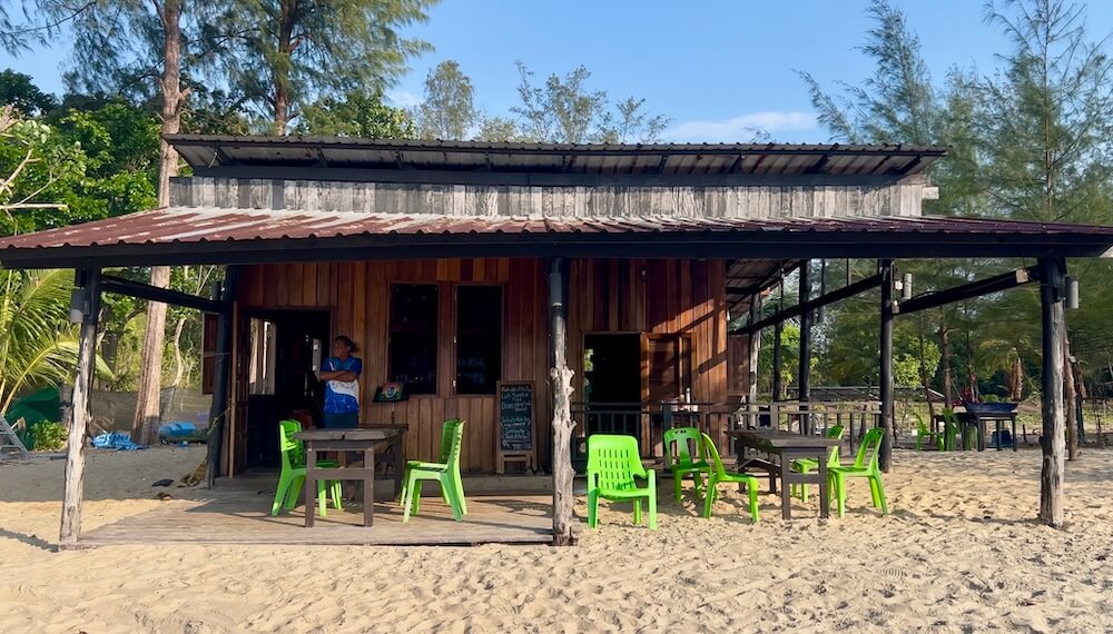 A rustic, wooden beach bar with a corrugated metal roof and lime green plastic chairs sitting on the sand at Koh Phra Thong.
