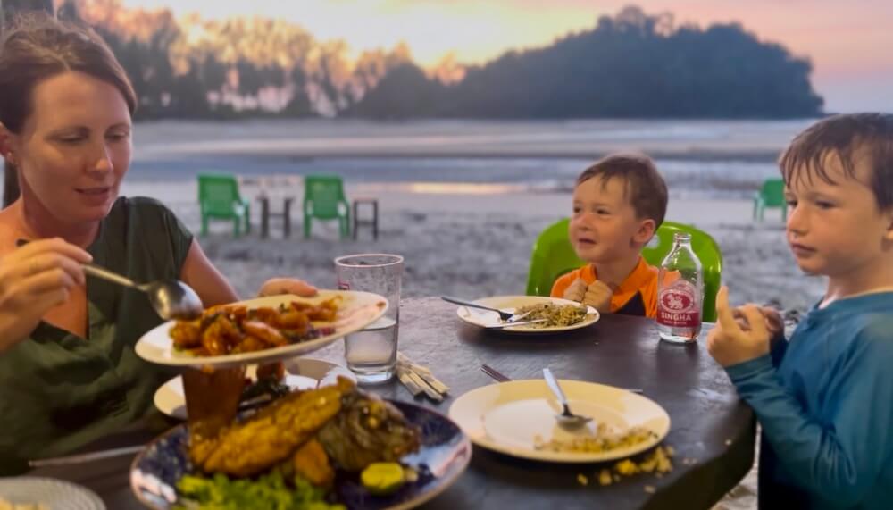 A woman and two young boys enjoying a fresh seafood dinner at a beach table during a vibrant sunset on Koh Phra Thong.