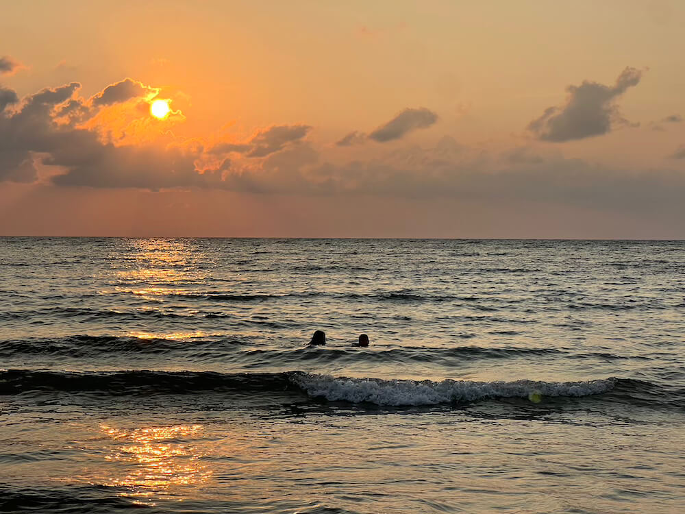 Two people swimming in the gentle waves of the Andaman Sea during a golden sunset with a glowing sun peeking through clouds.
