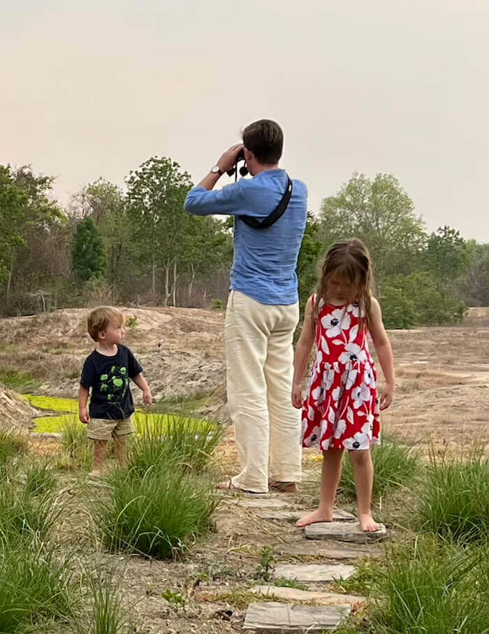 A man in a blue shirt uses binoculars to spot wildlife while two young children stand nearby on a stone path in a natural, open landscape.