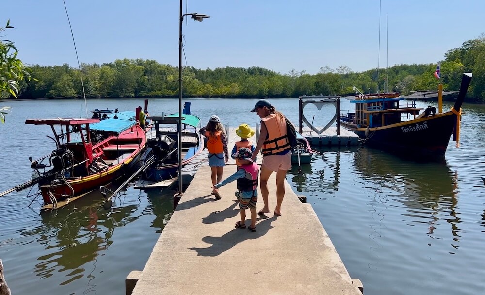 Setting Off from the Village A family wearing life jackets walks down a concrete pier toward several longtail boats tied up in a calm, mangrove-lined inlet.